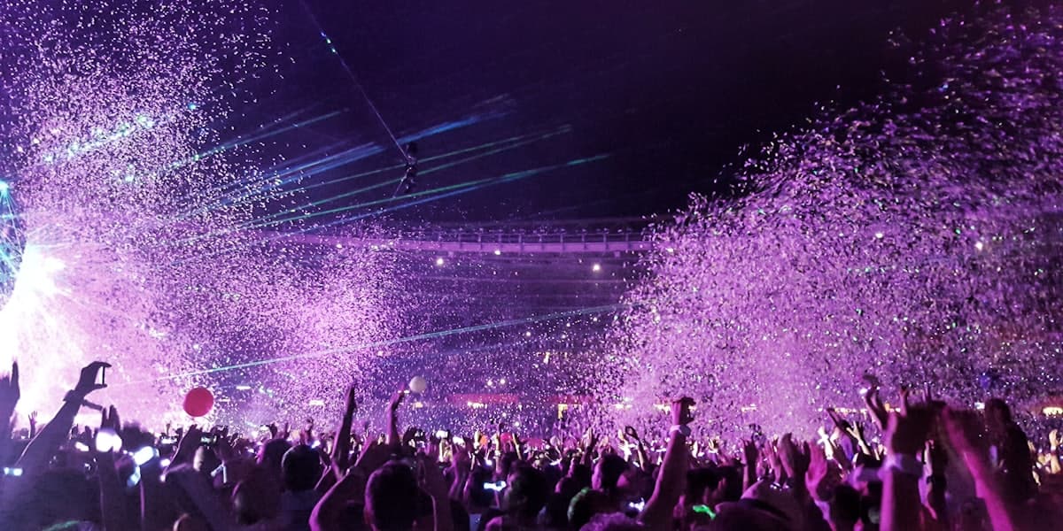 A packed arena crowd with hands raised under purple stage lights