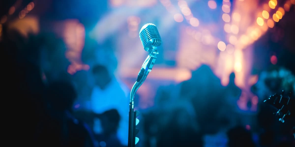 A vintage chrome stage microphone lit blue, a blurred live-music crowd and warm stage lights behind it