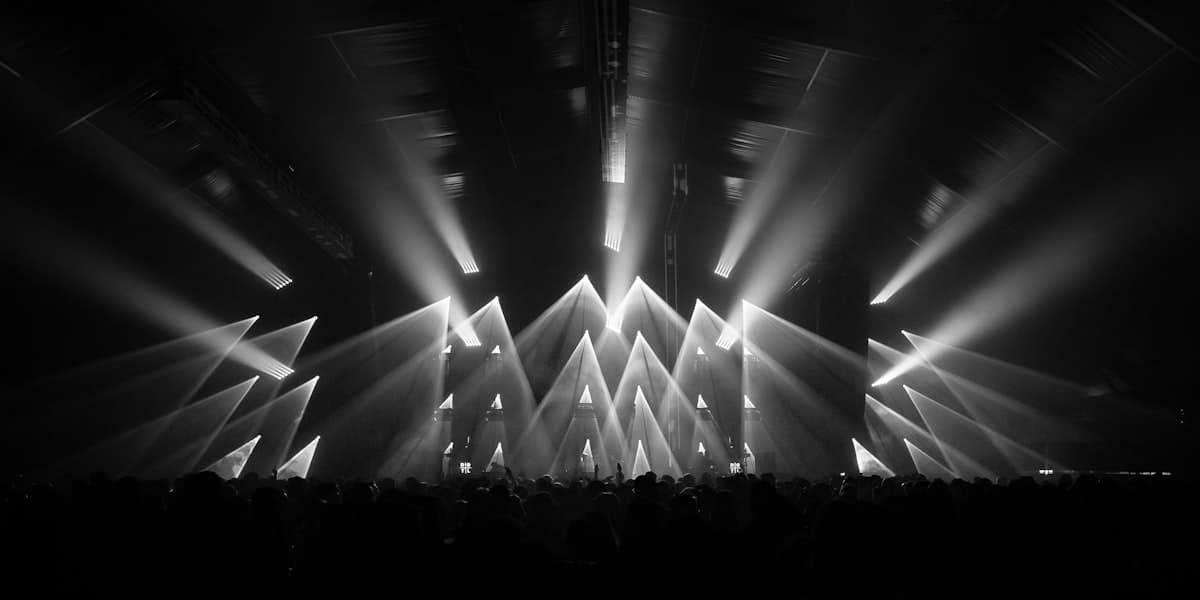 A full stage lighting rig casting beams over a concert crowd, in grayscale