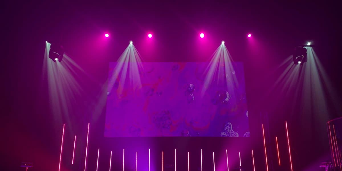 An empty stage lit magenta and white before doors — moving-head spots, a projection screen and rows of LED battens, with no performer and no crowd