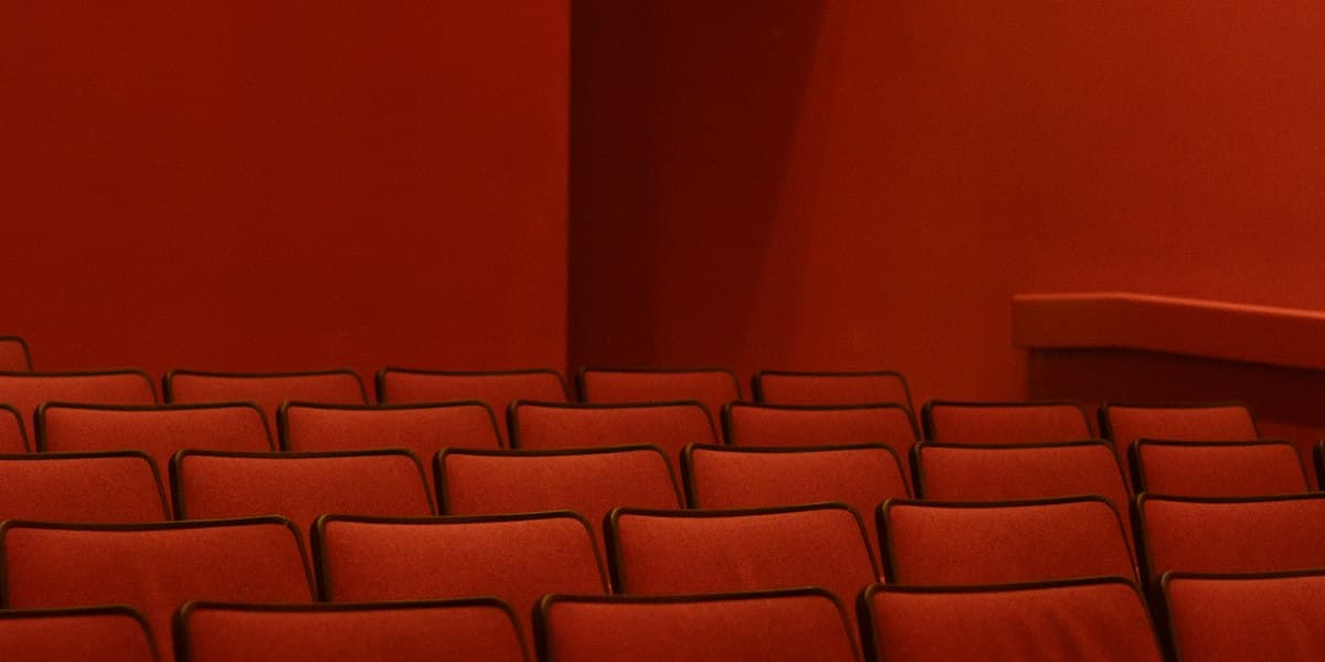 Rows of empty red upholstered seats in a house lit flat and low, angled toward a bare red wall — no stage, no signage and nobody in the room