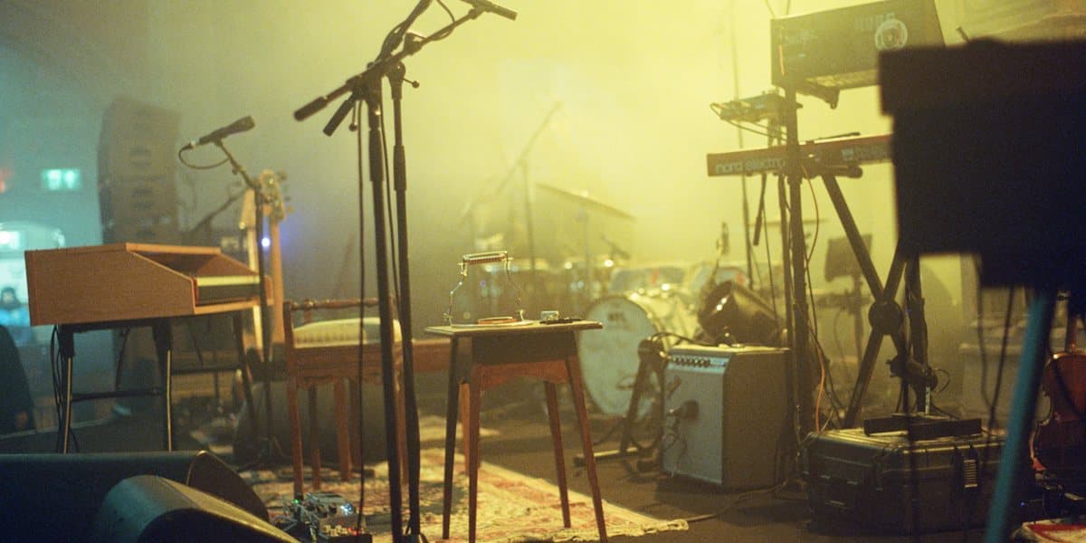 An empty stage set with backline — keyboards, a drum kit, an amplifier, mic stands and a road case — lit amber through haze before doors