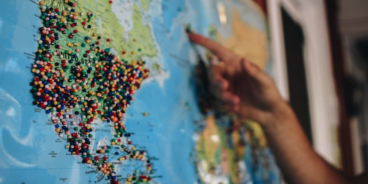 A person's hand pointing at a wall map with colored pins marking cities across the country