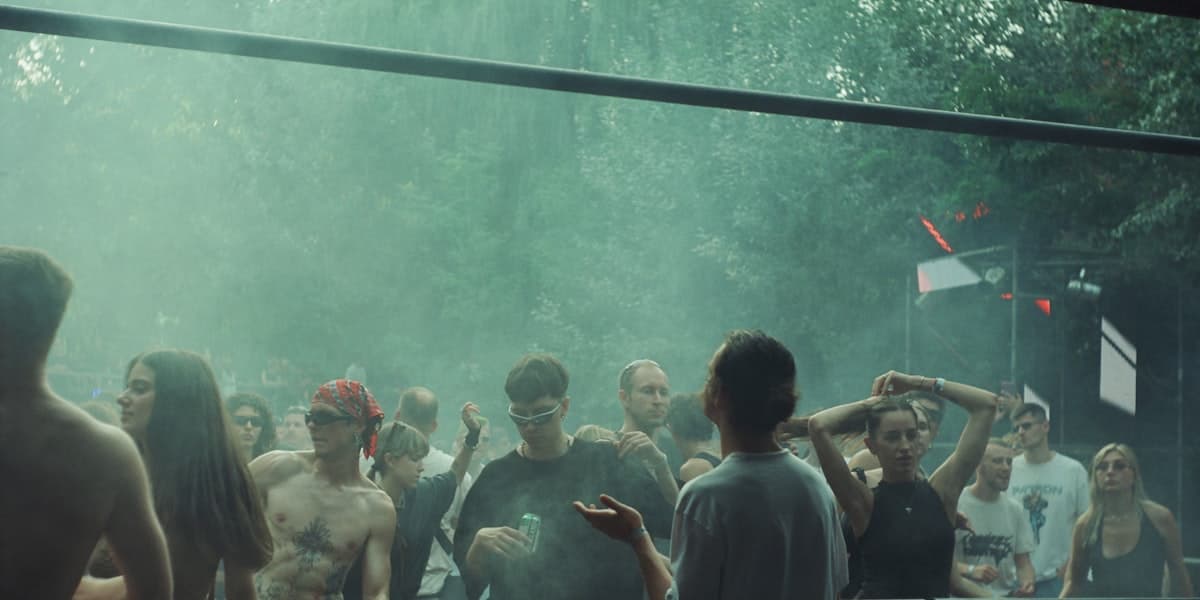 A crowd at an outdoor woodland music festival on a hazy summer day, some attendees shirtless in the heat